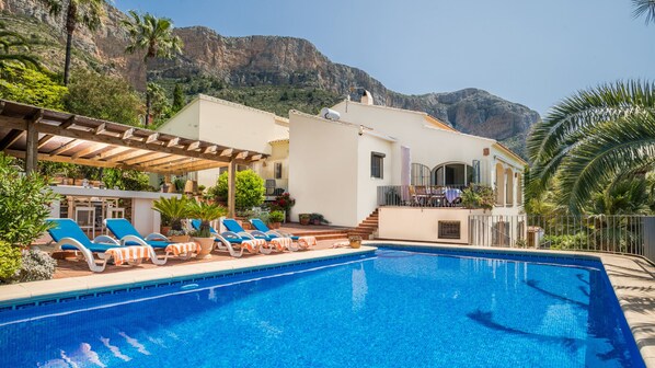 A swimming pool with lounge chairs, a pergola, and a mountain backdrop.