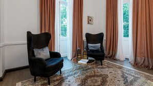 A room with two black armchairs, a patterned rug, a small round coffee table with books and decorative items, and large windows with brown curtains. - Superior Suite Theseus by 2 Mazi Suites (Athina)