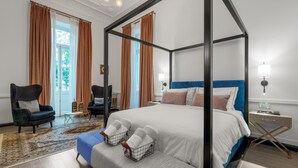 A four-poster bed with a blue headboard, a grey bench with white towels, a dark armchair, and a patterned rug. - Superior Suite Theseus by 2 Mazi Suites (Athina)