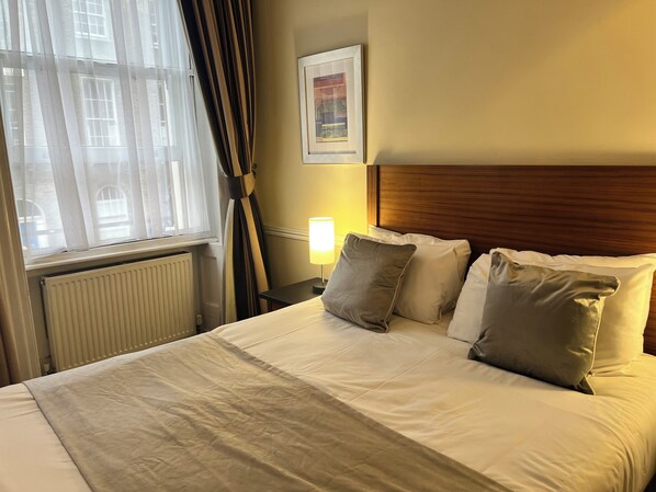 Quarto de hotel com uma cama grande, cabeceira de madeira, um quadro na parede e uma janela com cortinas.