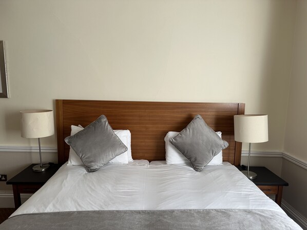 Un lit double avec une tête de lit en bois, deux oreillers gris, du linge de lit blanc et deux tables de chevet avec des lampes.