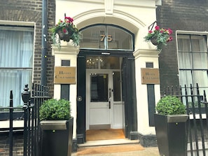 A entrada do Hotel Cavendish, com fachada em preto e branco, placa com o número 75 acima da porta e jardineiras com flores de cada lado.
