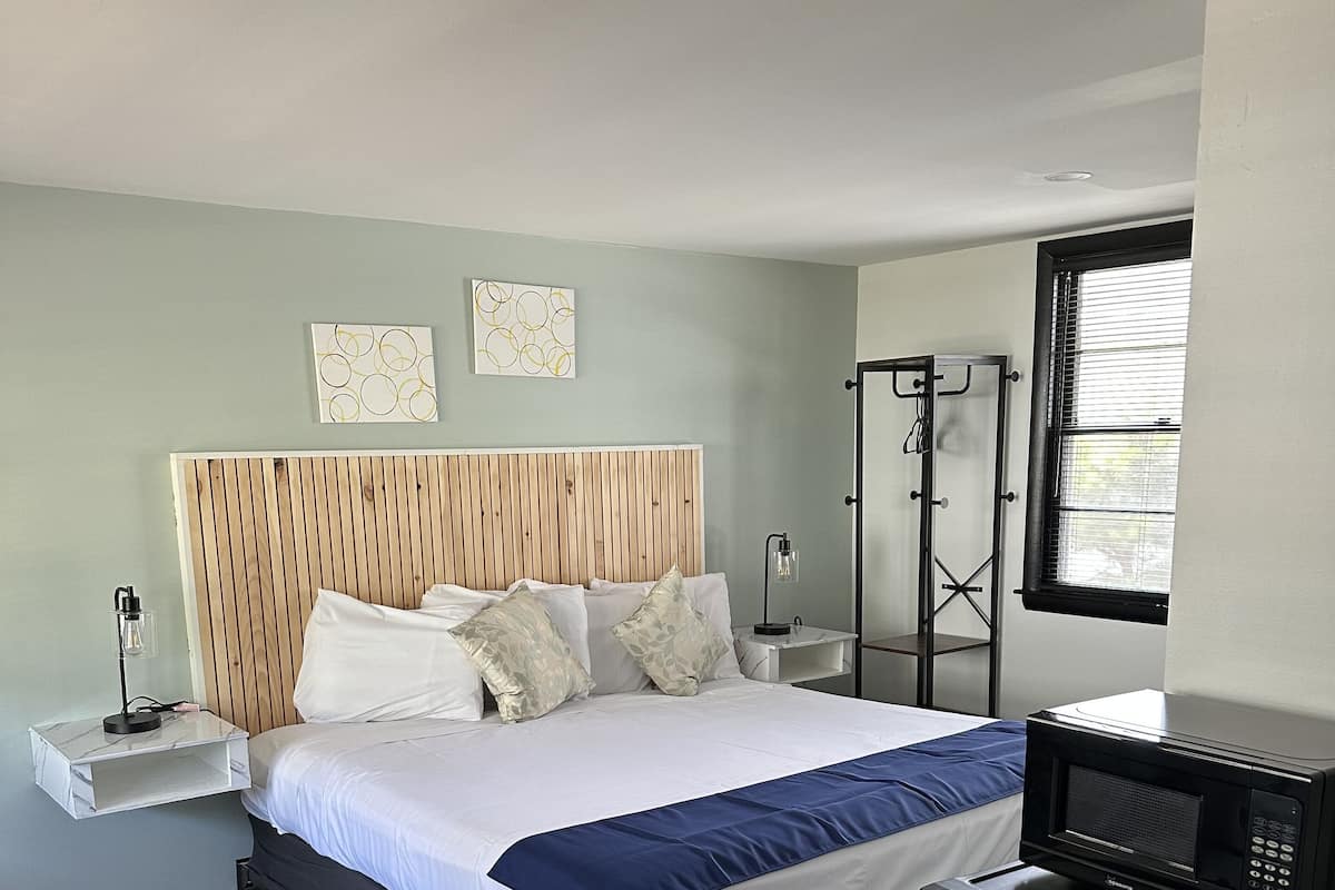 A modern hotel room with a wooden headboard, a bed with white and blue bedding, a small refrigerator, a bedside table with a lamp, and a window with blinds.