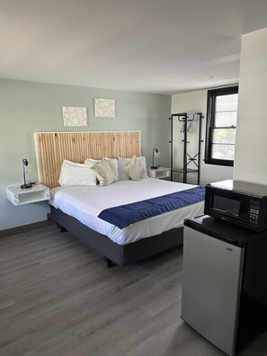Een moderne hotelkamer met een houten hoofdeinde, een bed met wit en blauw beddengoed, een kleine koelkast, een nachtkastje met een lamp en een raam met zonwering.