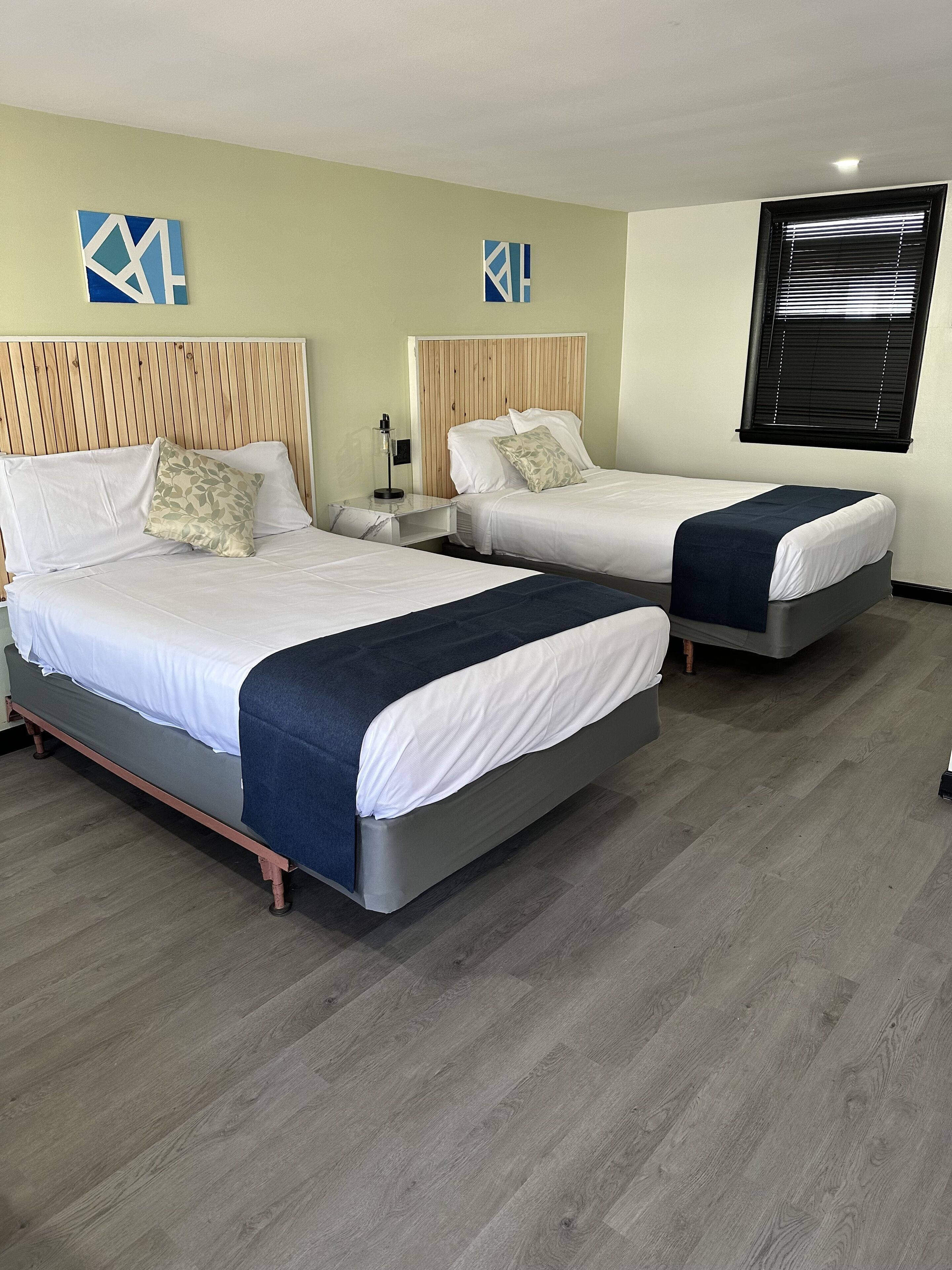 A hotel room with two beds, a wooden headboard, a nightstand, a lamp, and a window with blinds.