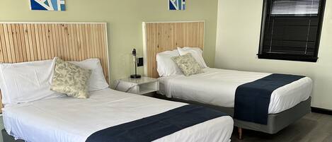 A hotel room with two beds, a wooden headboard, a nightstand, a lamp, and a window with blinds.