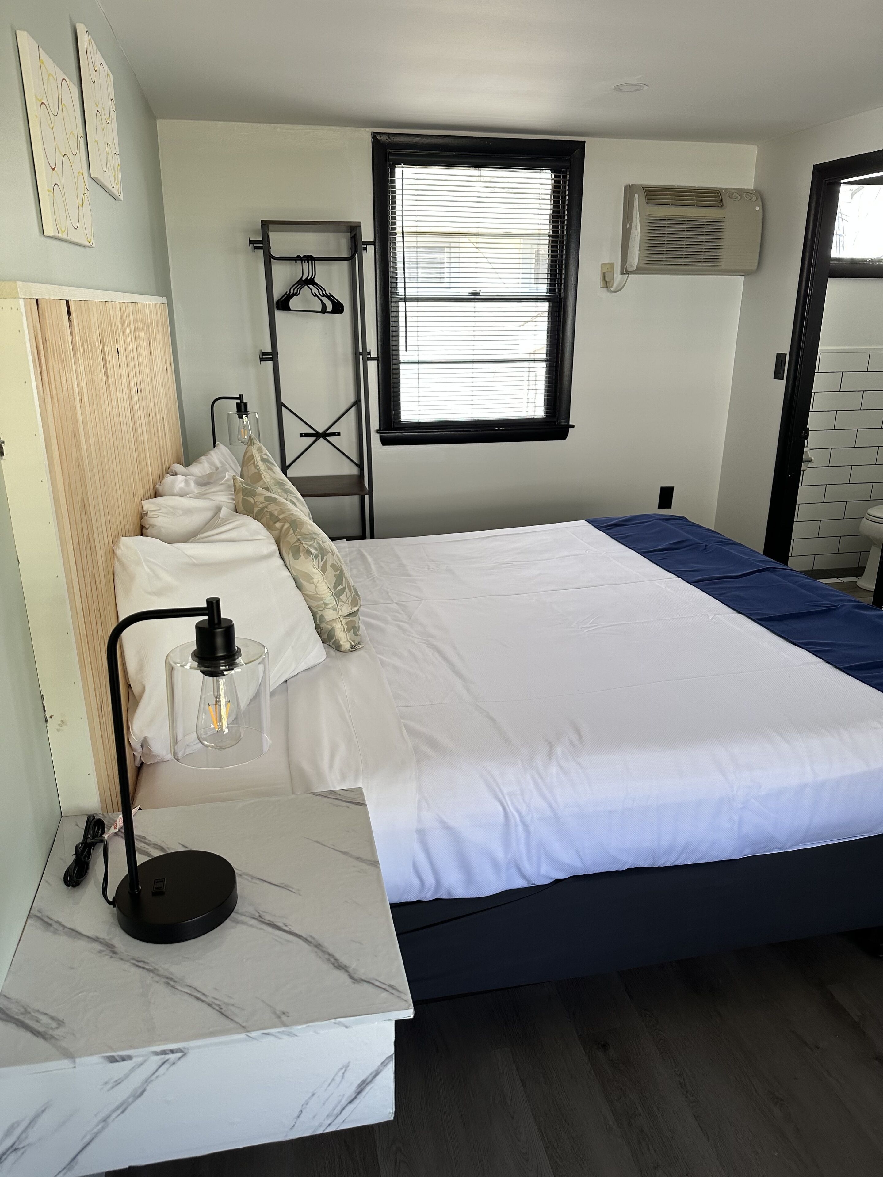 Un dormitorio con cama, mesita de noche con lámpara, ventana con persianas y aire acondicionado instalado en la pared.
