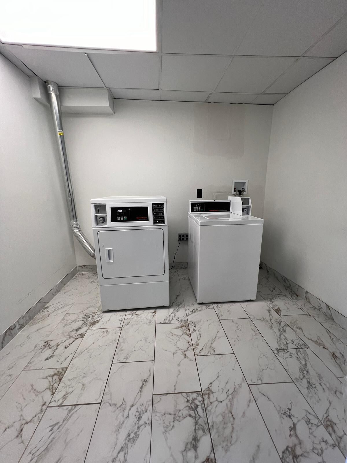 A laundry room with two washing machines and a dryer, marble-tiled floor, and a visible vent on the ceiling.