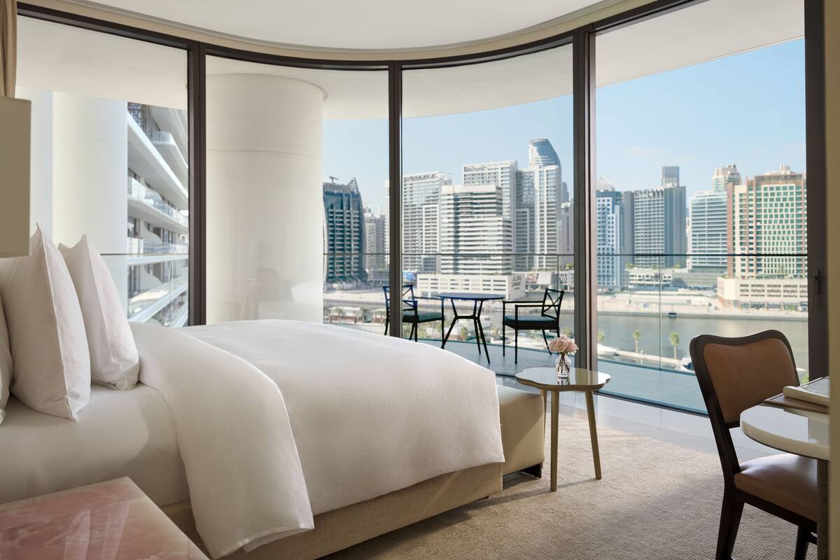 A modern hotel room with a large bed, a small table, and a view of the city skyline through large windows.