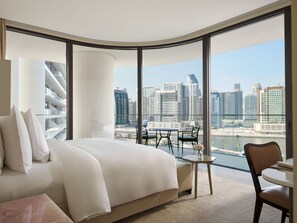 A modern hotel room with a large bed, a small table, and a view of the city skyline through large windows.