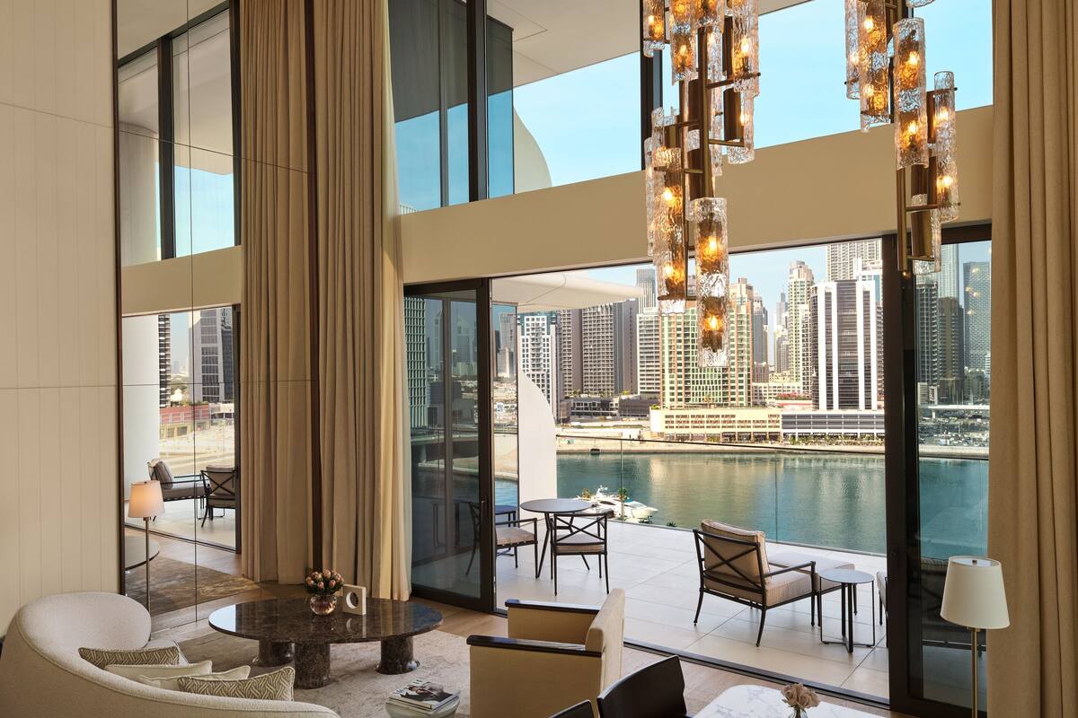 A modern hotel room with a large window overlooking a cityscape and water. It features a marble dining table, comfortable seating, and a unique chandelier.