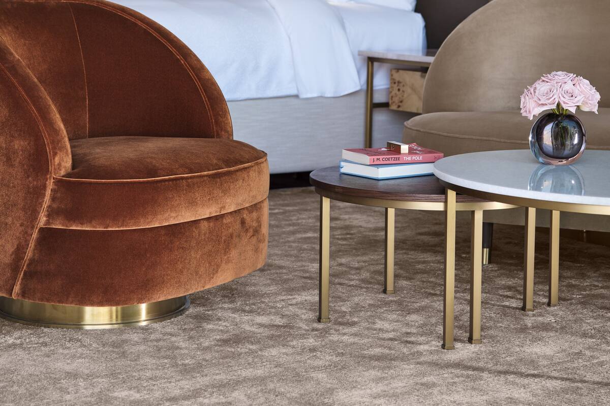 A hotel room with a large bed, a brown velvet armchair, a round coffee table, and a vase with flowers.