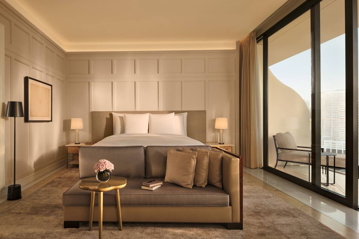 A modern hotel room with a large bed, a sofa, a small table, and a large window offering a city view.