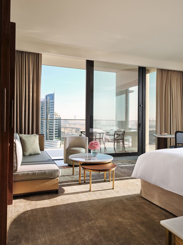 A hotel room with a large bed, a sofa, a small round table with a vase, a balcony with a chair, and a city view. - The Lana - Dorchester Collection (Dubai)