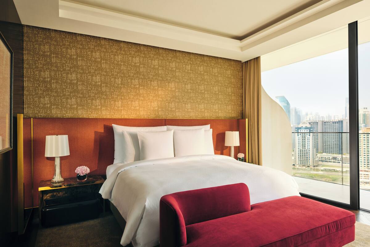 A hotel room with a large bed, a red bench, a nightstand with a lamp, and a view of a cityscape through large windows.