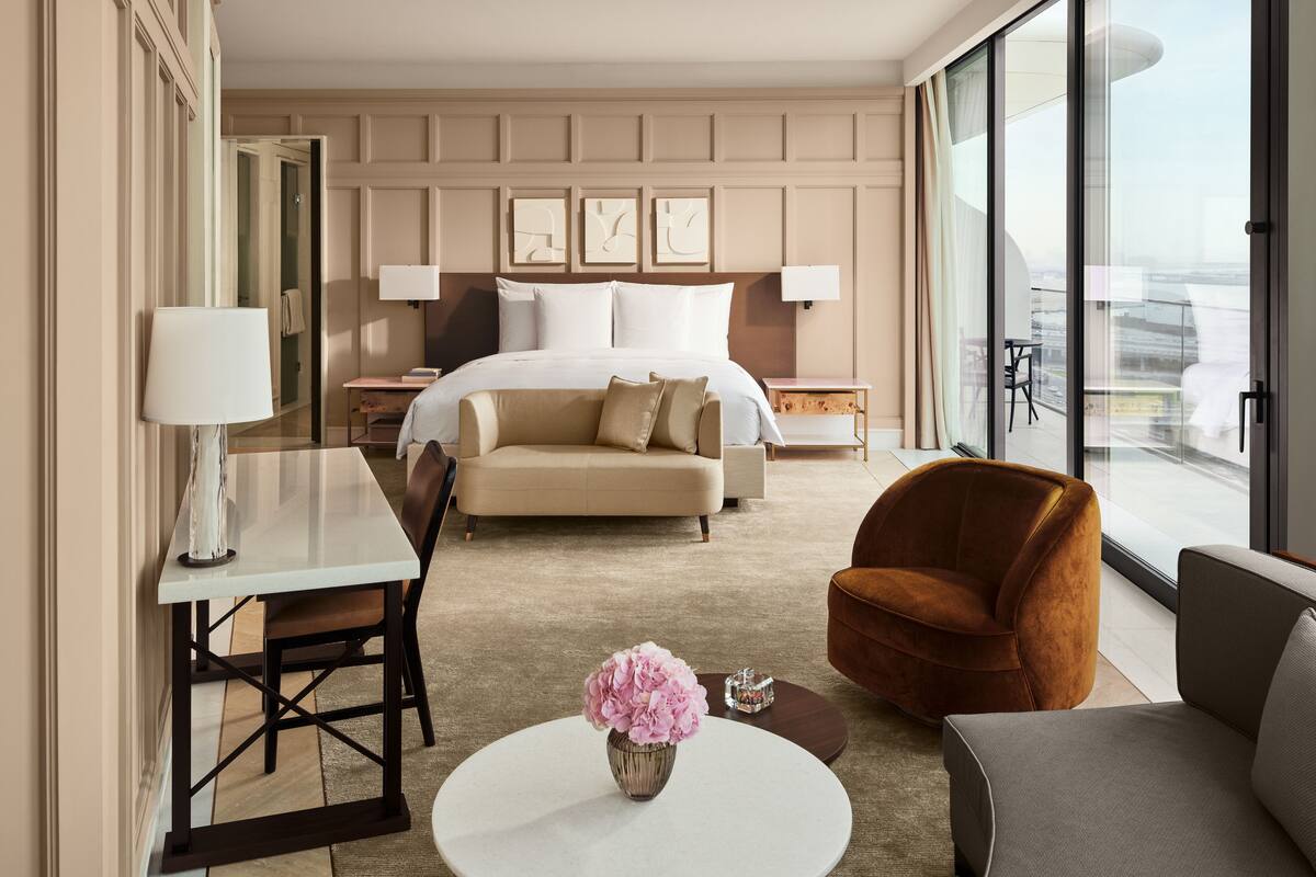 A hotel room with a large bed, a sofa, a chair, a small table, and a view of the city through large windows.