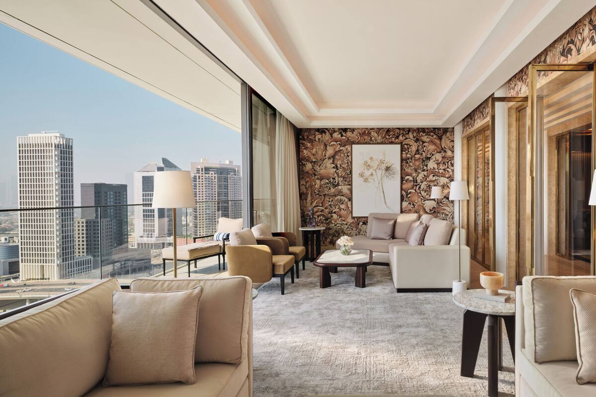 A modern hotel room with a city view, a cozy seating area with sofas and a coffee table, and a decorative wall with a floral pattern.