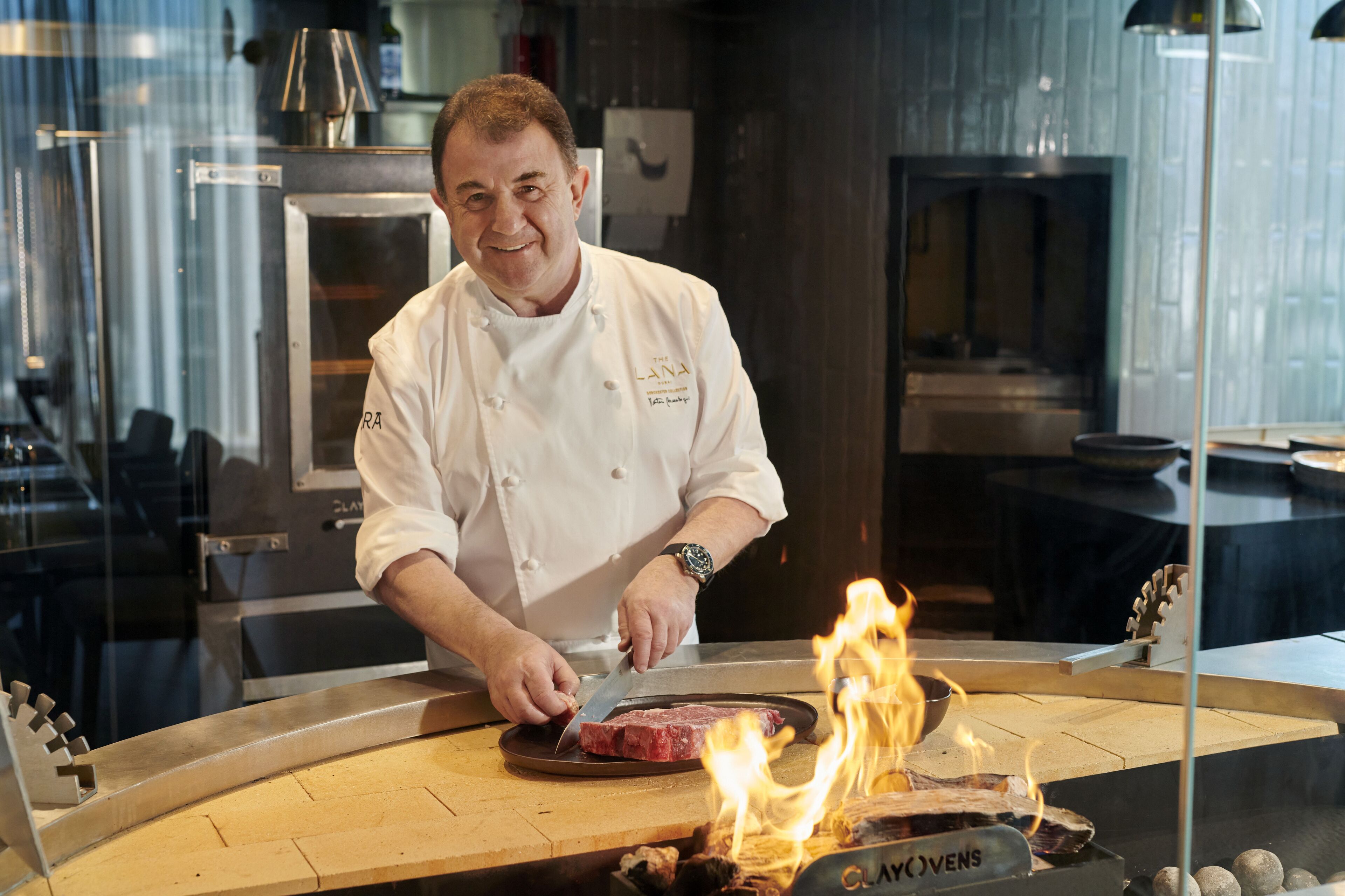 Un chef, vêtu d’un uniforme blanc, découpe de la viande sur une planche en bois, avec un gril visible au premier plan.
