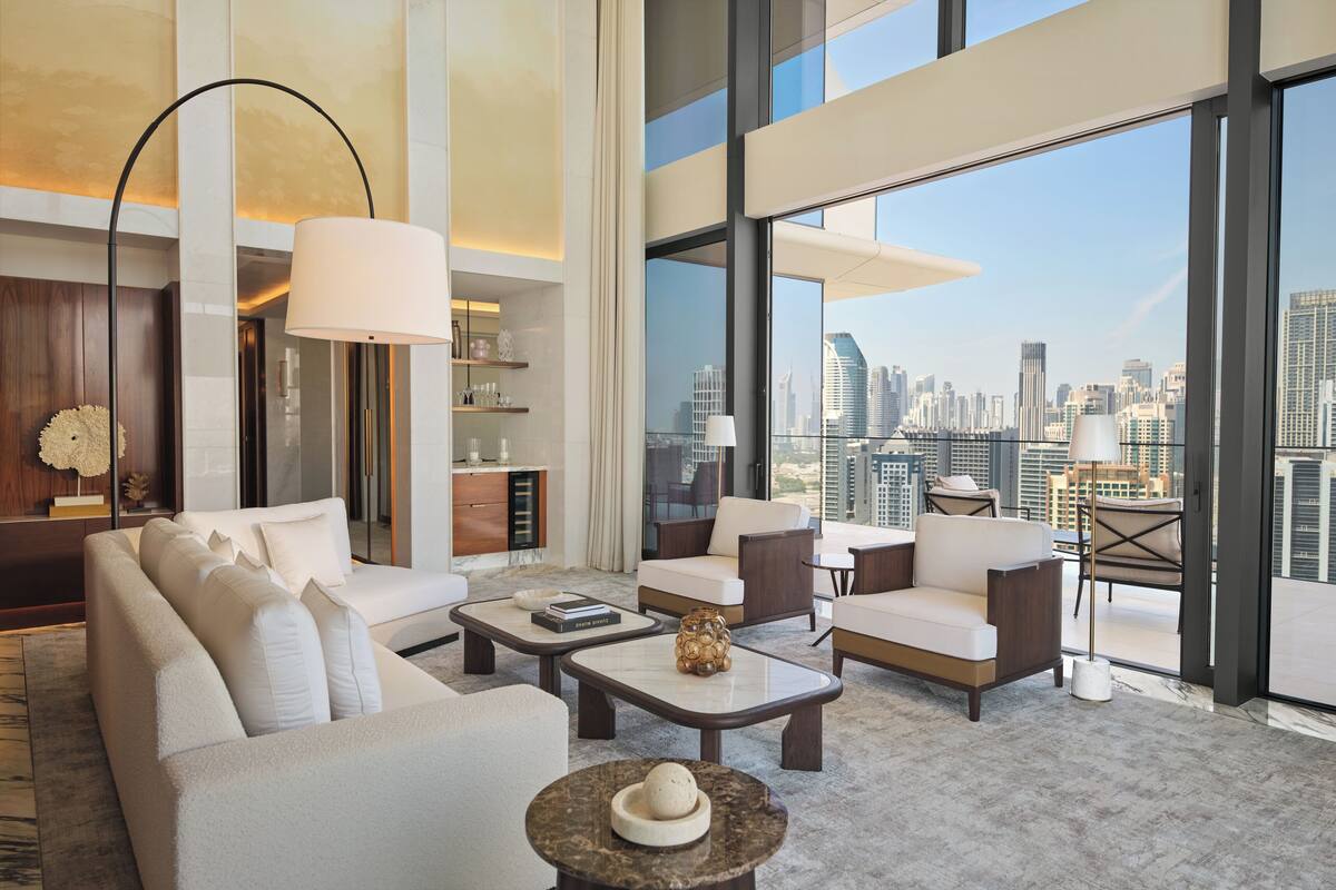A modern hotel room with a large city view, a sofa, armchairs, a coffee table, and a floor lamp.