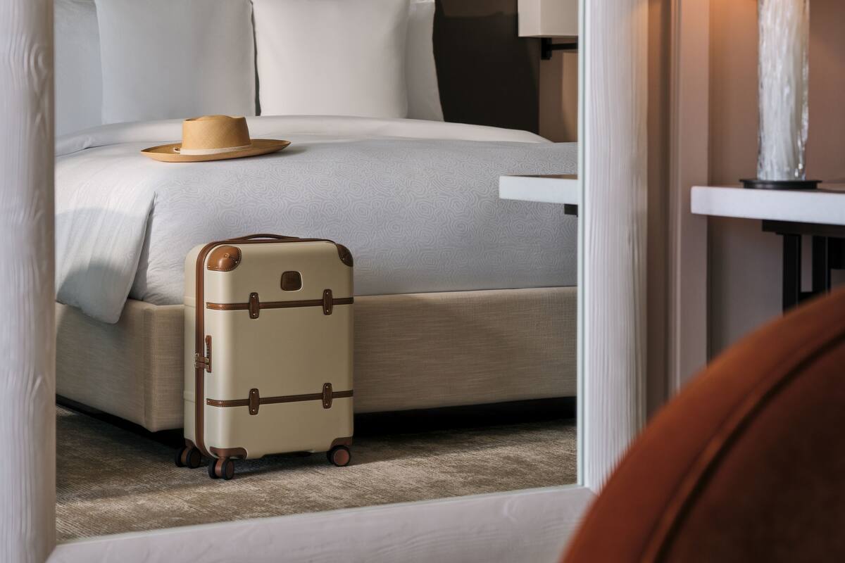 A neatly made bed with a suitcase and a hat on it, a bedside lamp, and a wall with paneling.