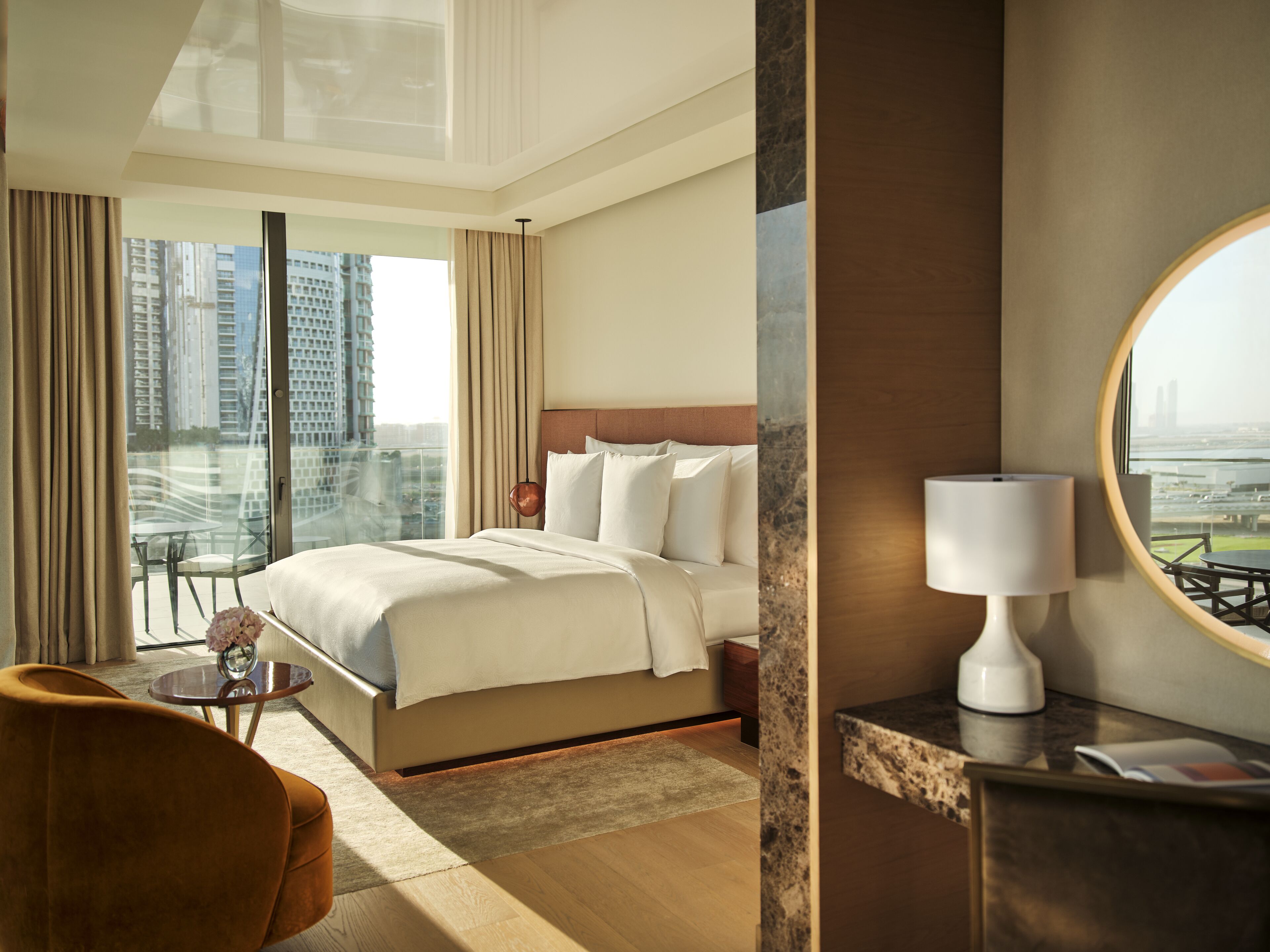 A hotel room with a large bed, a round mirror, a small table, and a view of the cityscape.
