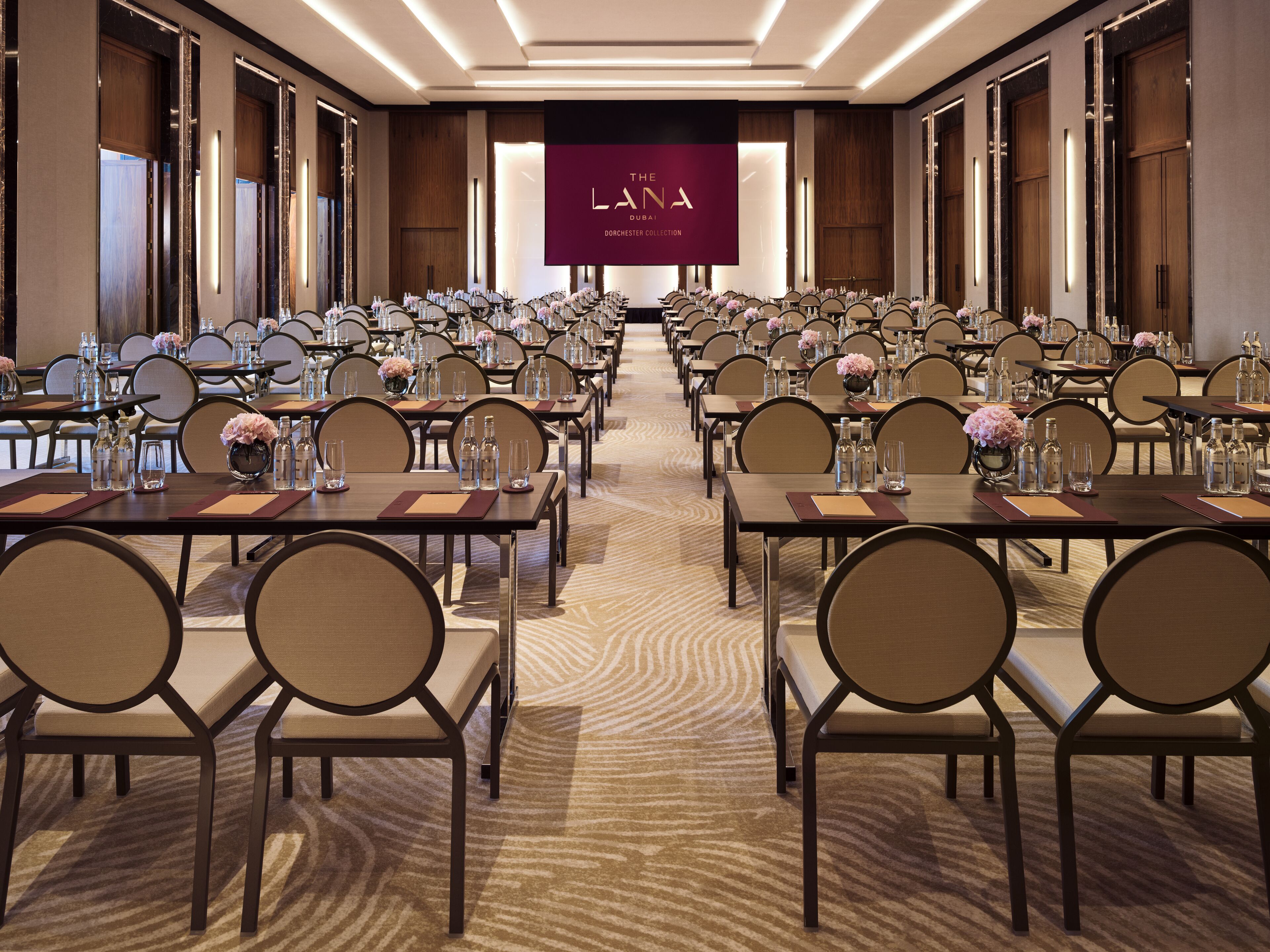 Un salón de conferencias con filas de mesas y sillas, una pantalla grande mostrando 'The Lana Dubai Dorchester Collection' y piso alfombrado.