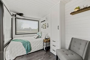 Ein Schlafzimmer mit Bett, Nachttisch, Sessel, Kleiderschrank, Fenster mit Jalousien und einem Regal mit Pflanze.