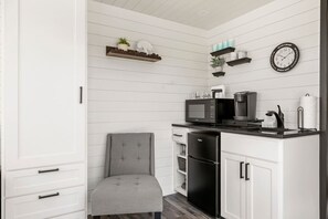 A compact kitchen with white cabinetry, a grey upholstered chair, a black refrigerator, a microwave, a coffee maker, and a wall clock.