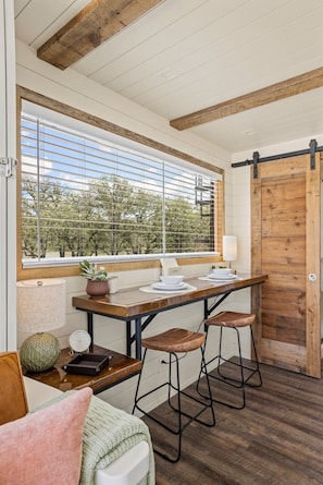 A kitchen with a wooden table, bar stools, a window with blinds, and a view of trees.