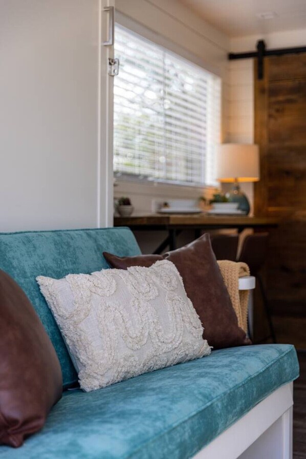 A teal sofa with patterned cushions, a wooden door, and a kitchen area in the background.