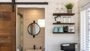 A bathroom with a white sink, a round mirror, and a coffee machine. There are shelves with glass jars and a potted plant. - New! The Desert Escape-Cozy Container Home (Fredericksburg)