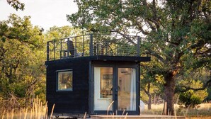 A small, dark-colored cabin with a balcony, surrounded by trees and grass. - New! The Desert Escape-Cozy Container Home (Fredericksburg)