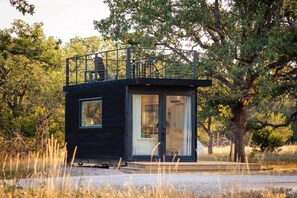 A small, dark-colored cabin with a balcony, surrounded by trees and grass.
