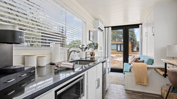 A modern kitchen with a black countertop, white cabinets, and a built-in oven. There is a window with blinds, a sink, and a view of the outside. - New! The Desert Escape-Cozy Container Home (Fredericksburg)