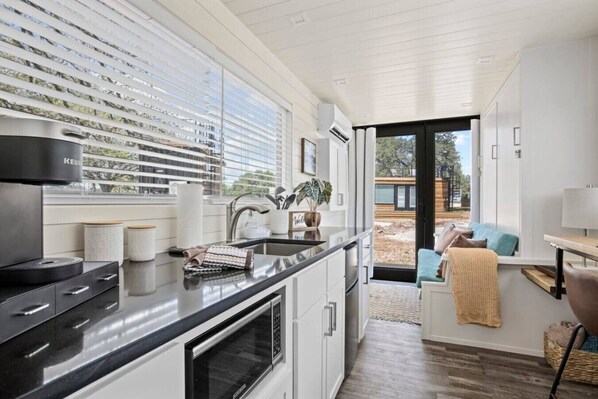 A modern kitchen with a black countertop, white cabinets, and a built-in oven. There is a window with blinds, a sink, and a view of the outside.