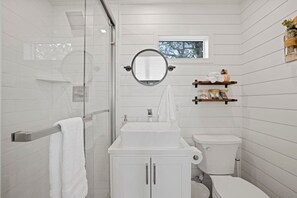 A modern bathroom with a glass shower enclosure, a white sink, a toilet, and a small window.