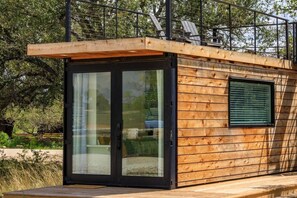 A modern wooden cabin with a sliding glass door entrance, a small window, and a deck. - New! Oak Grove-Container Retreat (Fredericksburg)