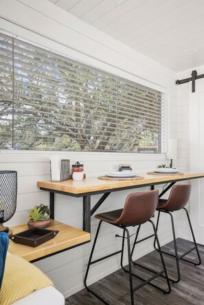 A modern kitchen with a large window, a wooden table, two bar stools, and a small plant on the counter.