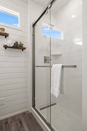 A modern bathroom with a glass shower enclosure, white tiled walls, a wooden shelf with towels, and a window above the shower. - The Texas Longhorn-Luxe CargoHome | Rooftop patio (Fredericksburg)