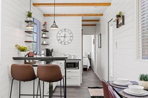 Une cuisine étroite aux murs recouverts de panneaux blancs, avec un plafond en bois, une horloge et un coin repas comprenant des chaises et une table.