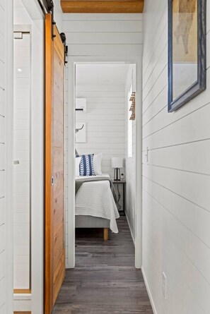 Um corredor estreito que dá para um quarto com cama, mesa de cabeceira e criado-mudo.