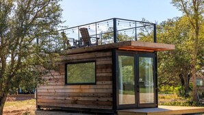 Una pequeña cabaña de madera con puerta de vidrio, baranda metálica en el nivel superior y terraza de madera.