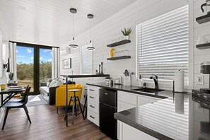 A modern kitchen with white cabinetry, a black countertop, and a dining area with a yellow chair. - New! The Yellow Beacon-Luxury Shipping Container (Fredericksburg)