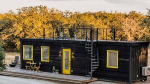 Eine kleine, schwarze Holzhütte mit gelber Tür und Fenstern, einer Treppe und Sitzgelegenheiten im Freien.