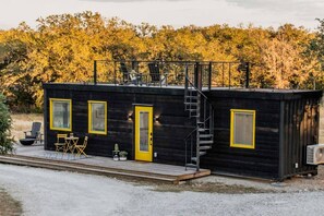 A small, black cabin with a yellow door and windows, a staircase, and outdoor seating. - New! The Yellow Beacon-Luxury Shipping Container (Fredericksburg)