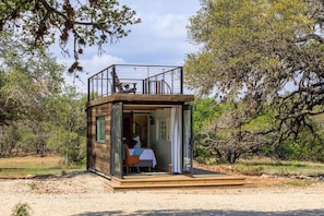 A small wooden cabin with a balcony, a bed, and a view of the surrounding trees.