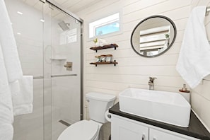 A modern bathroom with a glass shower enclosure, a white toilet, a large white sink, and a round mirror.