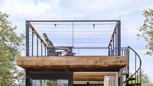 A compact, modern cabin with a glass door entrance, a small bedroom with a bed and a round mirror, and a wooden deck. - Container home | Roof Patio | Countryside Views (Fredericksburg)