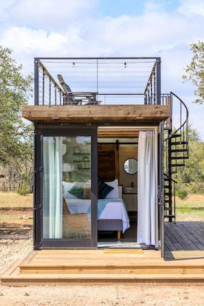 A compact, modern cabin with a glass door entrance, a small bedroom with a bed and a round mirror, and a wooden deck.