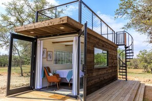 A cabin with a glass door entrance, a wooden deck, a spiral staircase, and a bed with a view. - Container home | Roof Patio | Countryside Views (Fredericksburg)